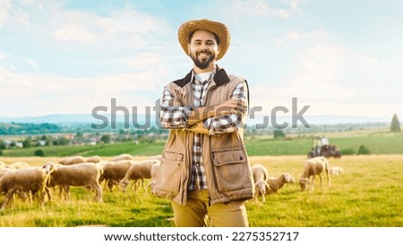 Portrait shot of handsome Caucasian cheerful man in motley shirt and hat standing in grasland, crossing hands and smiling to camera. Happy young male shepherd with sheep herd on background. Royalty-Free Stock Photo #2275352717