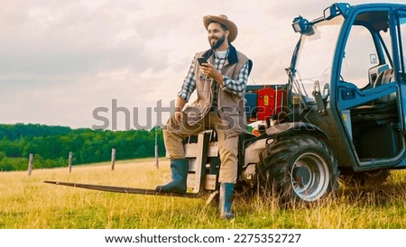 Caucasian handsome young man farmer in hat standing at tractor, using smartphone and resting in field. Countryside worker concept. Happy male having rest and texting on phone while chatting. Outdoor. Royalty-Free Stock Photo #2275352727