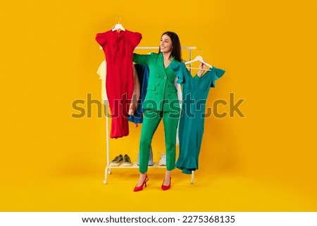 Shopaholic. Happy elegant woman choosing dresses holding two hangers, standing near clothing rail with trendy clothes over yellow background. Female fashion choice Royalty-Free Stock Photo #2275368135