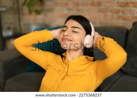 Happy mexican teen lady listening to music in wireless headphones, relaxing with hands behind head and eyes closed. Technology and relaxation concept