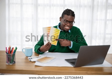 Cheerful African American Man Designer Choosing Color On Palette Showing It To Laptop Computer Video Calling Communicating And Working Online In Modern Office. Professional Design Concept