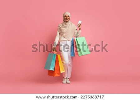 Smiling Muslim Woman In Hijab Walking With Smartphone And Shopping Bags In Hands, Arabic Shopaholic Lady Wearing Headscarf Enjoying App With Sales And Discount Offers, Pink Background, Copy Space Royalty-Free Stock Photo #2275368717