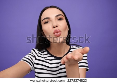 Beautiful young woman taking selfie while blowing kiss on purple background