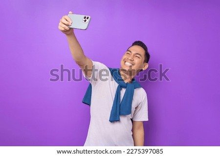 Cheerful young Asian man in white t-shirt doing selfie shot on mobile phone isolated on purple background