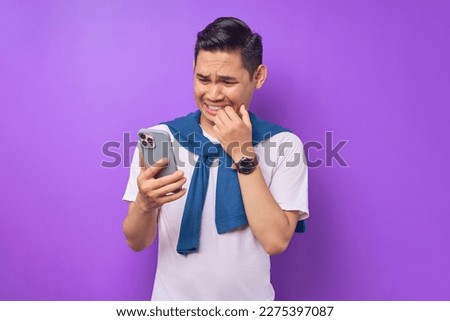 Scared young Asian man in casual t-shirt looking at mobile phone screen, reacting to bad news isolated on purple background. People lifestyle concept
