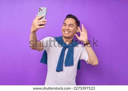 Cheerful young Asian man in white t-shirt doing selfie shot on mobile phone and waving hand isolated on purple background