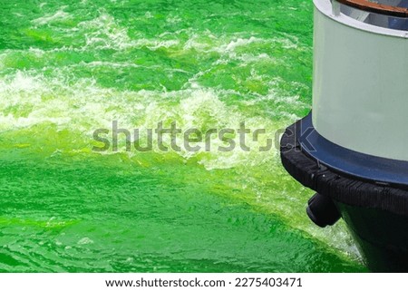 Close up photograph of a large barge boat moving through the bright green water of the Chicago River creating splashing waves and turbulence in wake during annual St. Patrick's day celebration event.