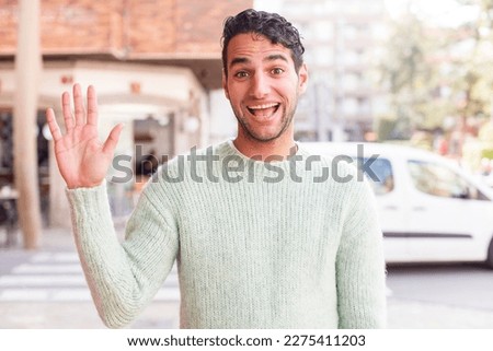 young hispanic man smiling happily and cheerfully, waving hand, welcoming and greeting you, or saying goodbye Royalty-Free Stock Photo #2275411203