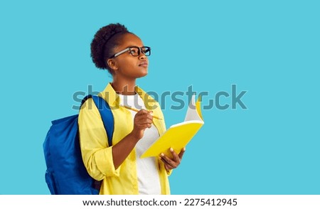 Portrait happy beautiful African American student girl in yellow shirt and eyeglasses standing isolated on blue background, holding pencil and notebook, writing essay, looking up and thinking of ideas Royalty-Free Stock Photo #2275412945
