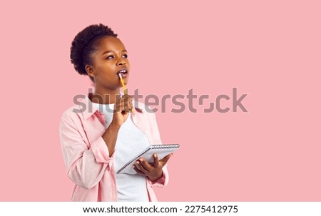 Beautiful African American woman in casual shirt standing on blank pink background, holding notebook, looking away, biting pencil and thinking. Female student or copywriter writing ideas for new Royalty-Free Stock Photo #2275412975