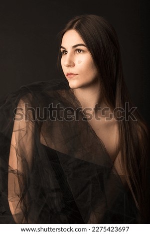 Fashion portrait of a beautiful caucasian model in her 20s. She is wrapped in tulle fabric. The background is black and she is posing. The model has brown eyes and long hair. 