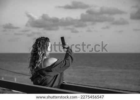 a beautiful blonde woman with long wavy hair stands on a bridge and shoots a sunset at the sea on her phone, black and white photo