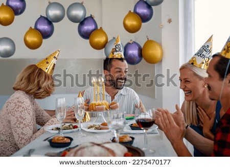 Smiling Multiethnic friends at Birthday dinner party laughing and enjoying together. Mid Adults with party hats and balloons have fun on Birthday lunch indoors