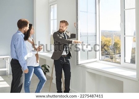 Real estate agent shows new home. Male realtor shows apartment for sale and its view from window to young married couple. Family looking for house to buy listens to man in suit. Real estate concept. Royalty-Free Stock Photo #2275487411