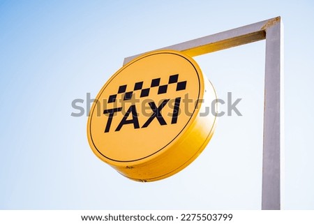 A taxi sign hangs against the sky, a taxi stop, a yellow banner with black text, letters in a circle, taxi checkers, sunlight. High quality photo