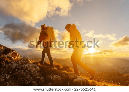 couple of two hikers, man and woman, together with their dog, ascending a mountain peak at sunset with the sun in the background. sport and adventure. Weekend activities. Royalty-Free Stock Photo #2275525641