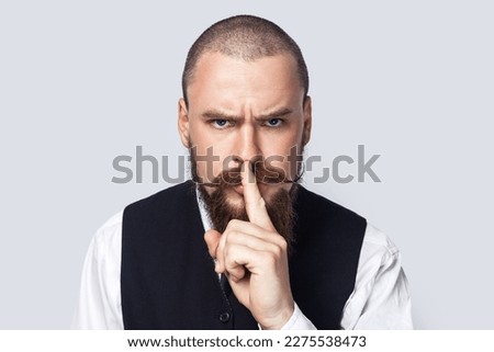 Keep silence. Portrait of serious strict man with beard and mustache standing and showing shh gesture, looking at camera with bossy expression. Indoor studio shot isolated on gray background.