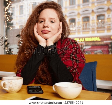 Young red-haired girl with coffee and a smartphone in a cafe, the girl chats in instant messengers during a coffee break Royalty-Free Stock Photo #2275542041