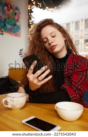 Young red-haired girl with coffee and a smartphone in a cafe, the girl chats in instant messengers during a coffee break Royalty-Free Stock Photo #2275542043