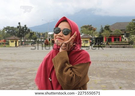 Asian woman in a red hijab smiling and wearing sunglasses with a mountain view as a background on Dieng Wonosobo Indonesia Tourism