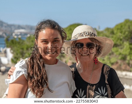 Grandmother and her granddaughter on a summer day posing for a family photo. Grandma wears a hat and red earrings. The granddaughter a white shirt.