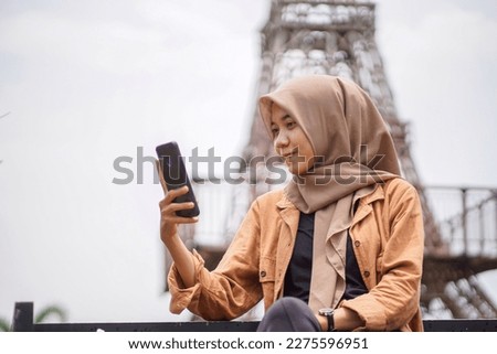 Asian woman wearing modern hijab playing social media using her smartphone in park and eiffel tower replica background