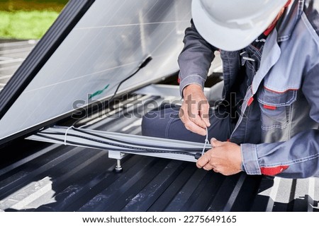 Close up view of male worker securing cables by special tie. Solar battery installation on roof of house. Man engineer in white helmet securing all cables of solar PV panel together. Royalty-Free Stock Photo #2275649165