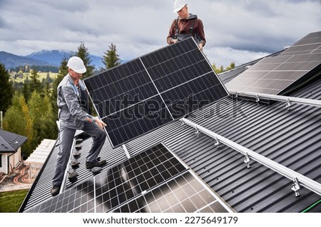 Engineers building solar panel system on roof of house. Men workers in helmets carrying photovoltaic solar module outdoors. Concept of alternative and renewable energy. Royalty-Free Stock Photo #2275649179
