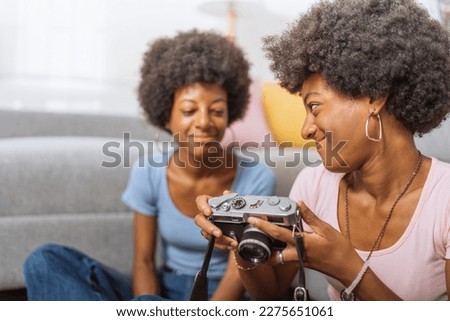 Twins with afro hairstyle and jeans, looking at the pictures they took with the old camera, sitting on the living room floor of their apartment.