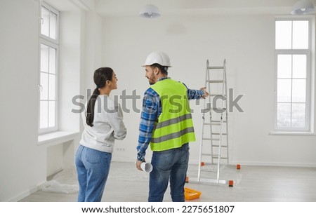 Architect, engineer or foreman in a uniform and hard hat standing at home in a room with a step ladder and talking to a young woman about house construction