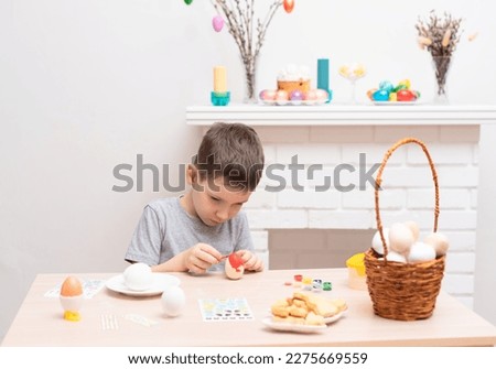 Child boy painting easter eggs against defocused fireplace with Easter decorations on it. Selective focus. Happy Easter concept. Process of crafting at home