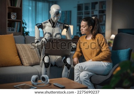 In the living room, a young woman and her humanoid robot companion use a laptop to learn, showcasing the potential of AI. Royalty-Free Stock Photo #2275690237