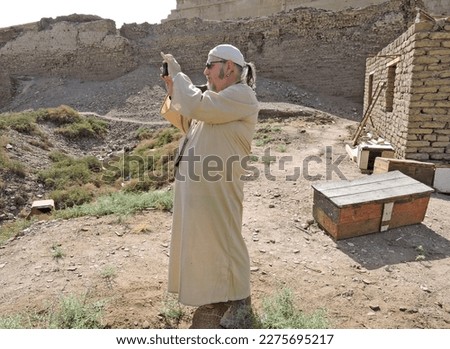 European senior man is taking a picture with his smartphone in Dendera, Egypt. He is wearing a beanie, an Egyptian Jellabiya and military boots. #uniqueSSelf