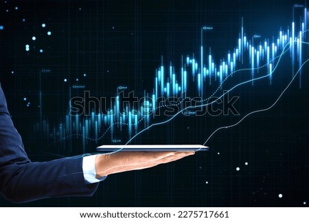 Investing and online trading concept with digital tablet on businessman hand on dark technological background with blue growing financial chart candlestick and diagram