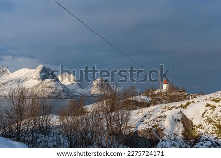 lighthouse in the fjord  of Husøy on the island Senja, Norway. Øyfjorden, steep snowy mountain sides rise out of the ocean, winter time. beacon for ships with white base and red roof, standing on rock