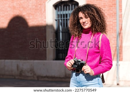 Young curly haired woman in casual outfit standing on town street against brick building with vintage photo camera while exploring city