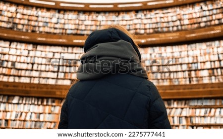 A girl in a scarf and a top stands with her back in the Stockholm Public Library against the backdrop of several floors of books