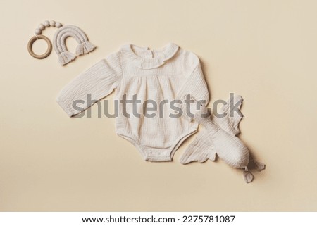 Mockup of cream infant bodysuit made of organic cotton with eco friendly baby accessories, knitted rainbow, soft duck on beige background. Gift for newborn baby. Top view. Flat lay. Copy space.