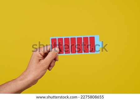 a man is holding a cardboard depiction of a fully charged battery against on a yellow background Royalty-Free Stock Photo #2275808655