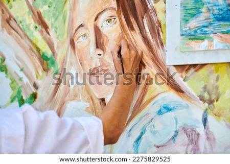 Girl artist hand holds paintbrush and draws surreal woman portrait on white canvas at outdoor art painting festival, paintings art picture process. Woman artist paints atmospheric surreal picture