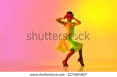 Music in movements. Emotional little girl in yellow stage dress dancing classical ballroom dance over gradient pink-yellow background in neon light filter. Concept of beauty, professional dances Royalty-Free Stock Photo #2275845407