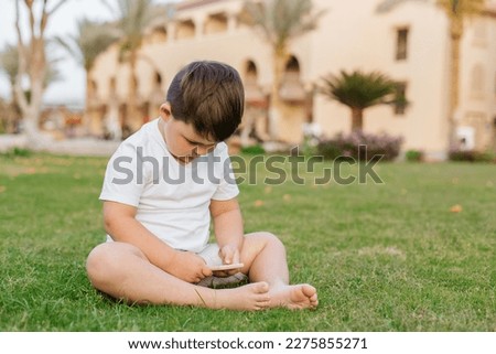 Little cute curious baby boy with blond curly hair in light summer clothes sitting on lawn with fresh green grass sunny day outdoor holding mobile phone, horizontal picture