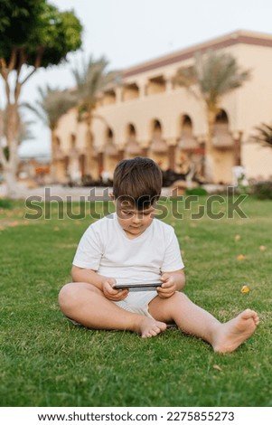 Little cute curious baby boy with blond curly hair in light summer clothes sitting on lawn with fresh green grass sunny day outdoor holding mobile phone, horizontal picture