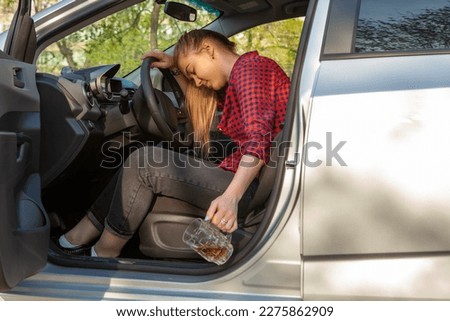 Drunk young girl driving a car with a mug of beer on sunset background, dangerous driving concept.