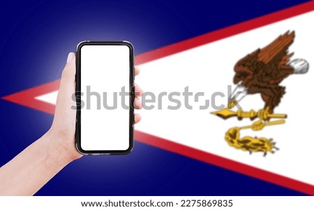 Male hand holding smartphone with blank on screen, on background of blurred flag of American Samoa. Close-up view.