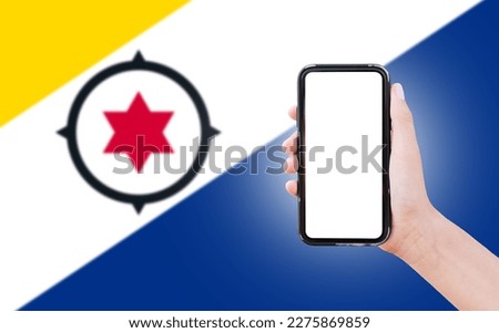 Male hand holding smartphone with blank on screen, on background of blurred flag of Bonaire. Close-up view.