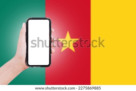 Male hand holding smartphone with blank on screen, on background of blurred flag of Cameroon. Close-up view.