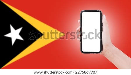 Male hand holding smartphone with blank on screen, on background of blurred flag of East Timor. Close-up view.