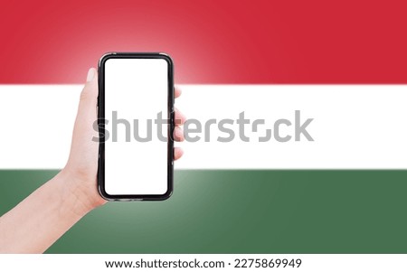 Male hand holding smartphone with blank on screen, on background of blurred flag of Hungary. Close-up view.