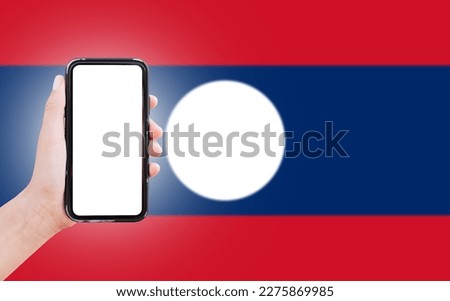Male hand holding smartphone with blank on screen, on background of blurred flag of Laos. Close-up view.